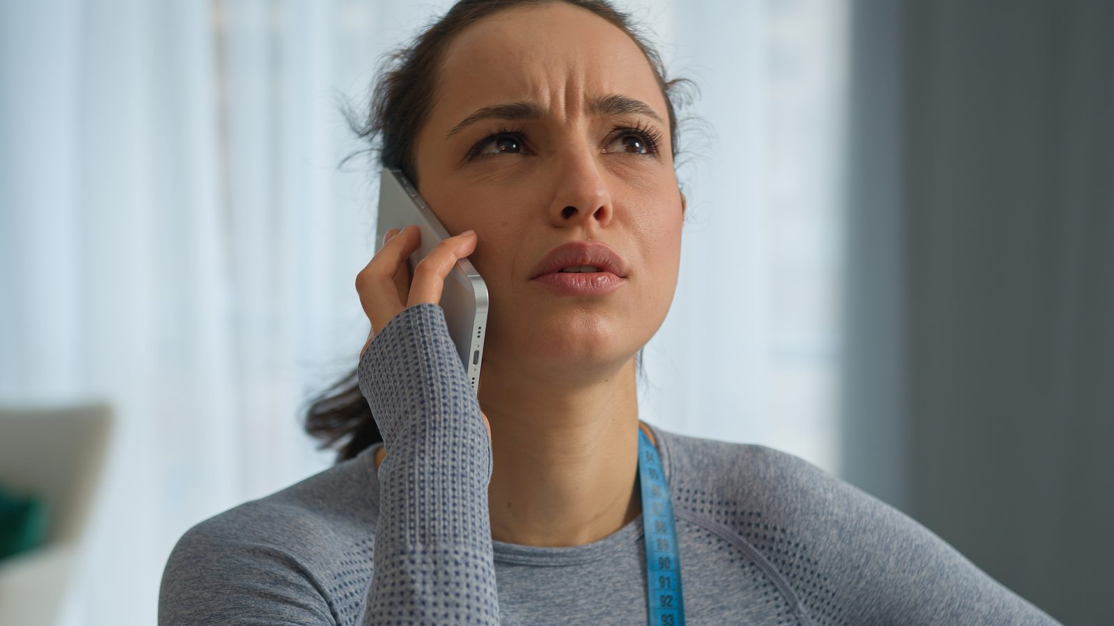 A woman in a grey top holds a phone to her ear and looks worried.