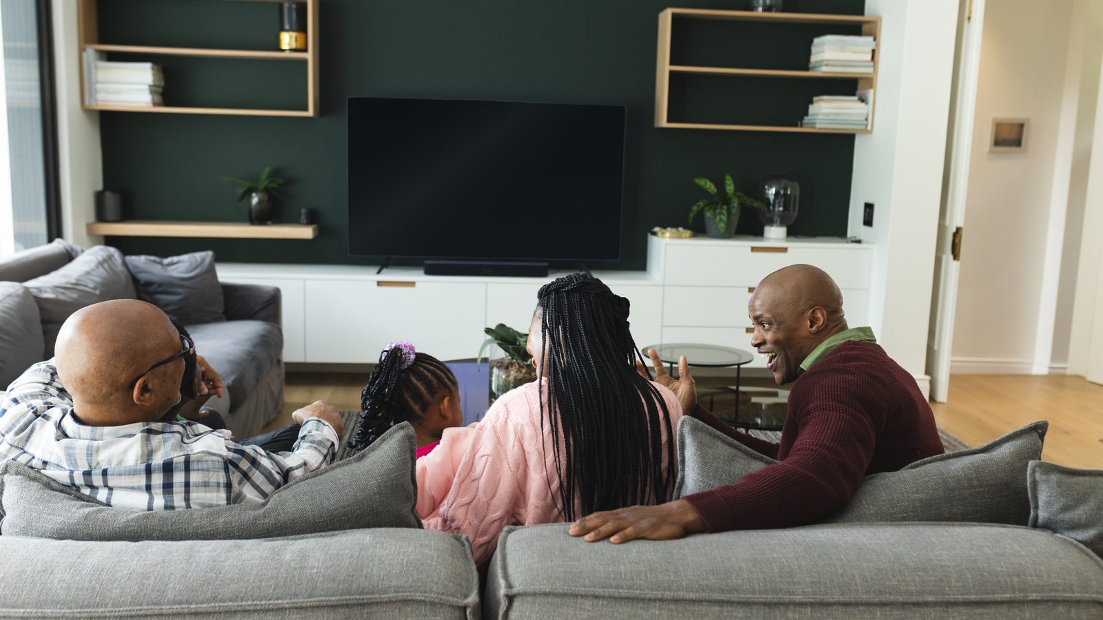 A family sits together on a gray sofa in a living room, facing a large flat-screen TV that is switched off.