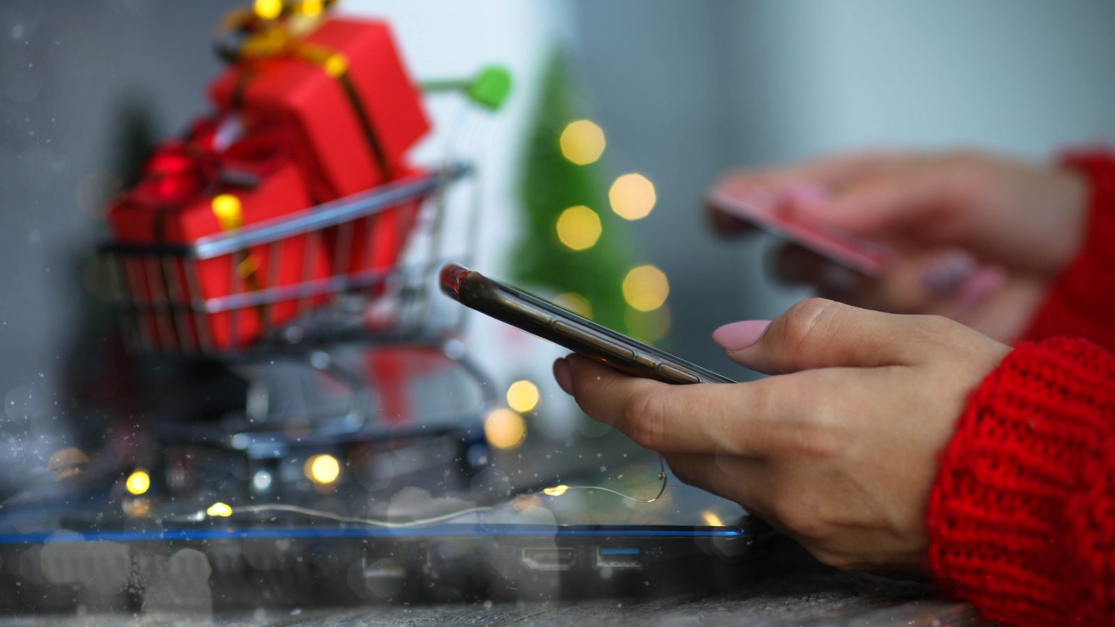 A person in a red sweater shops on a phone next to a small cart full of wrapped red gifts and holiday lights.