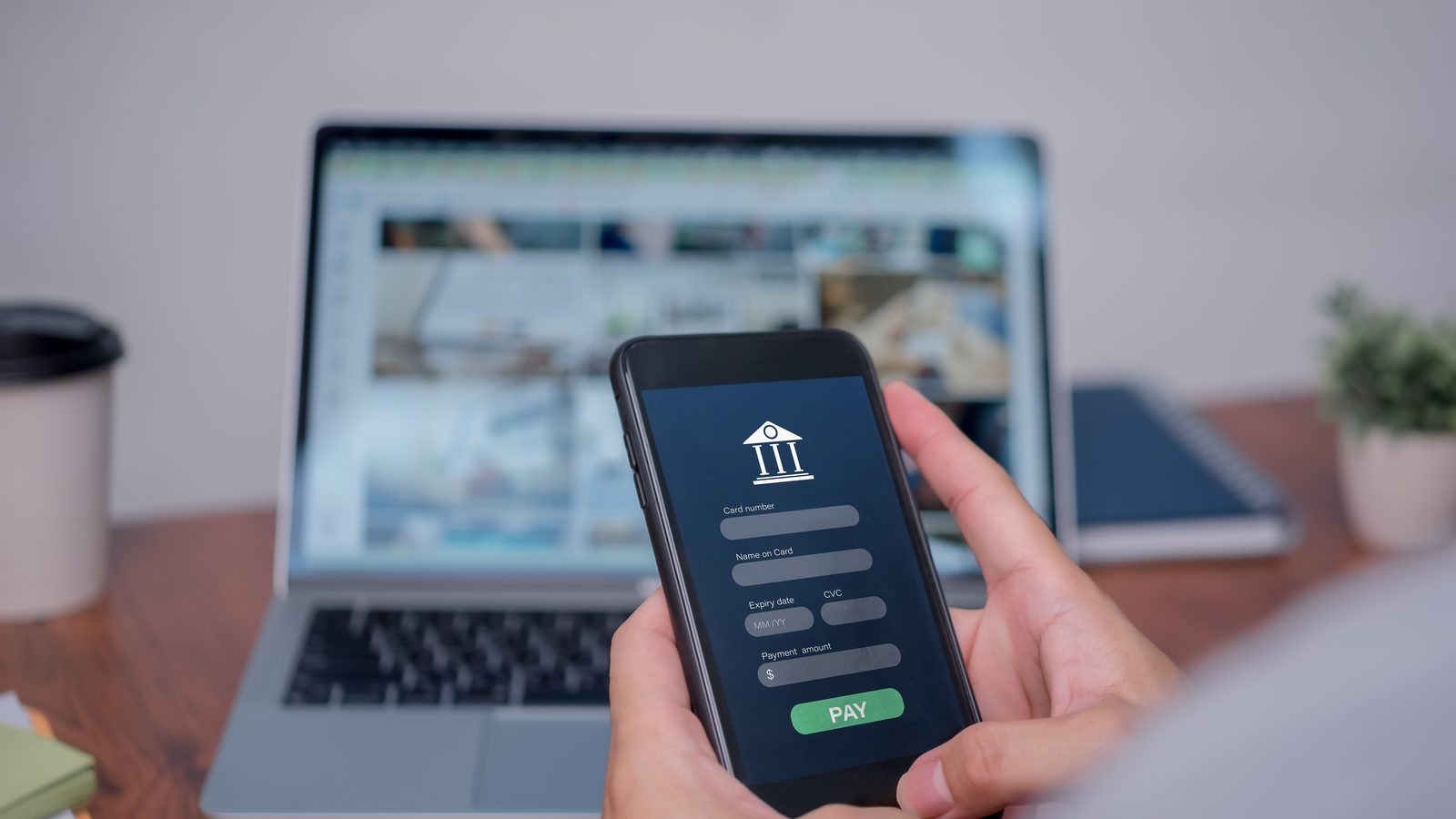 Hands hold a phone showing a banking app payment screen with a green Pay button, in front of a laptop.
