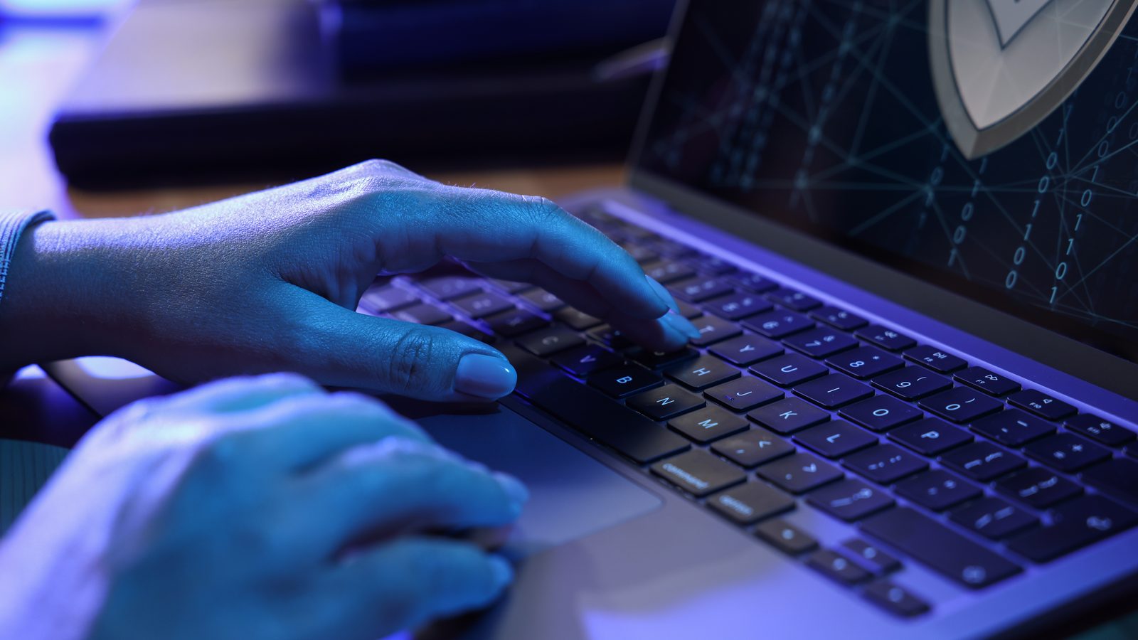 Hands type on a laptop lit in blue, with a security shield graphic on the screen.