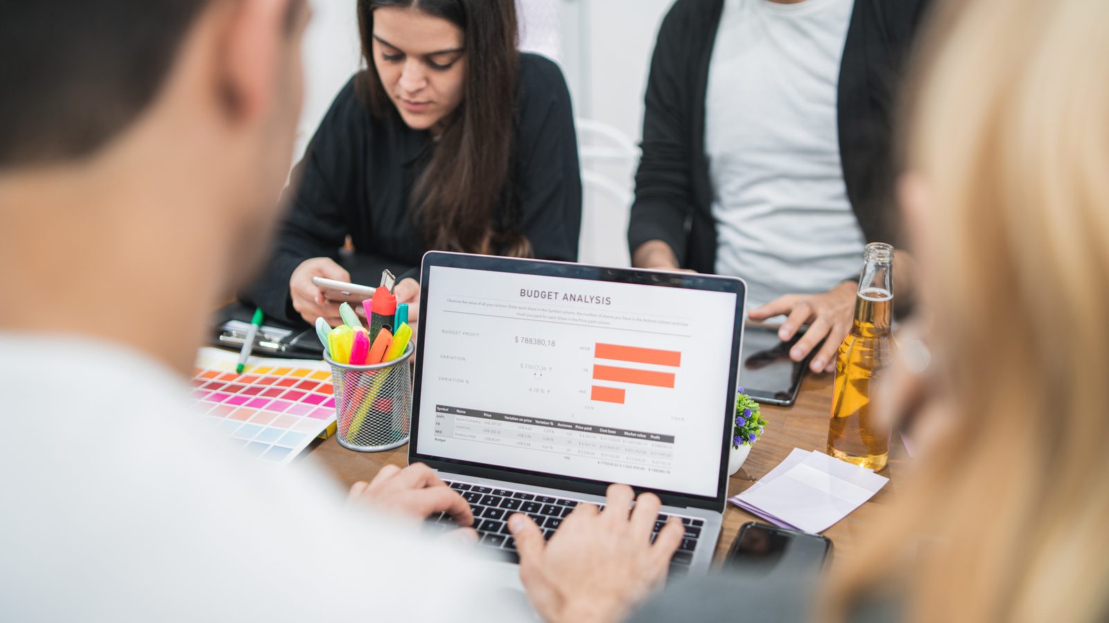 Four coworkers gather around a laptop on a cluttered desk, its screen showing a slide titled Budget Analysis with an orange bar chart.