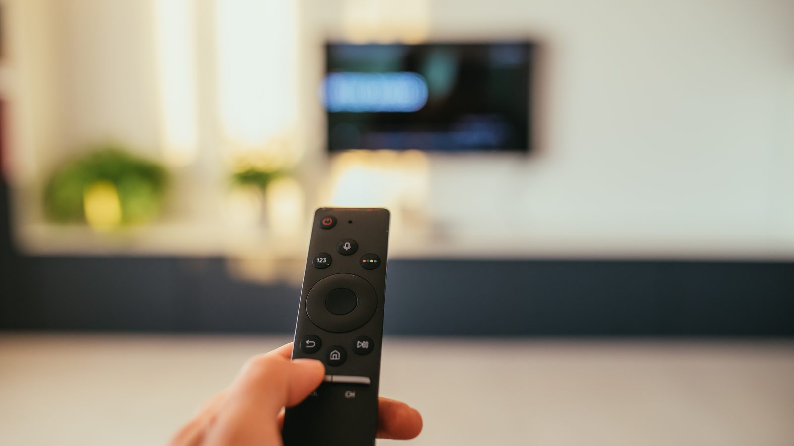 A hand points a black TV remote toward a blurred flat-screen TV mounted on a living room wall.