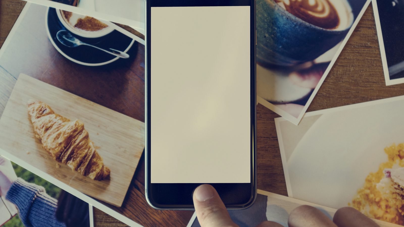 A finger taps a blank phone screen lying on a scatter of printed photos of coffee and food.