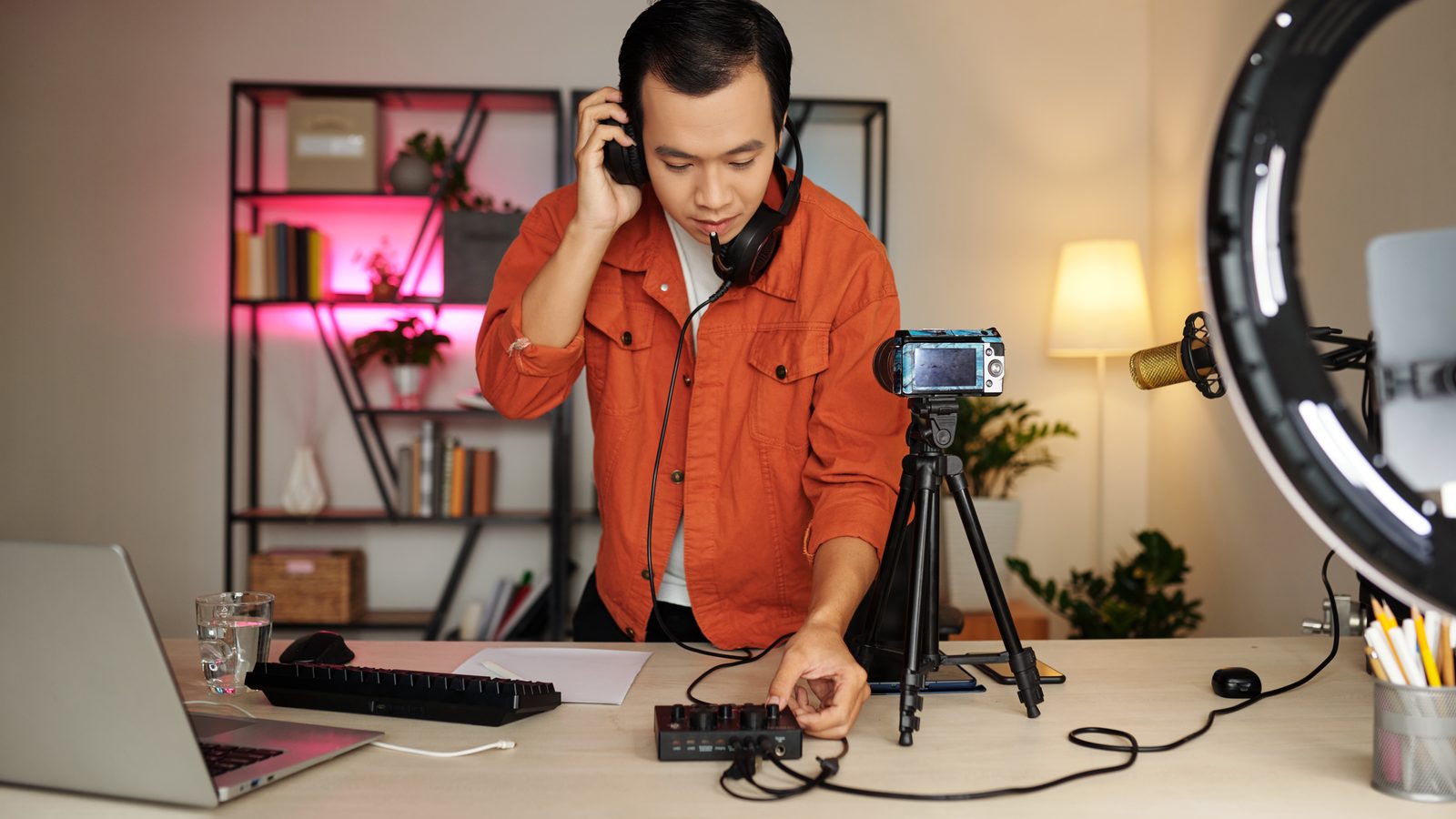 A young man in headphones adjusts a small mixer at a home studio desk with a camera and ring light.