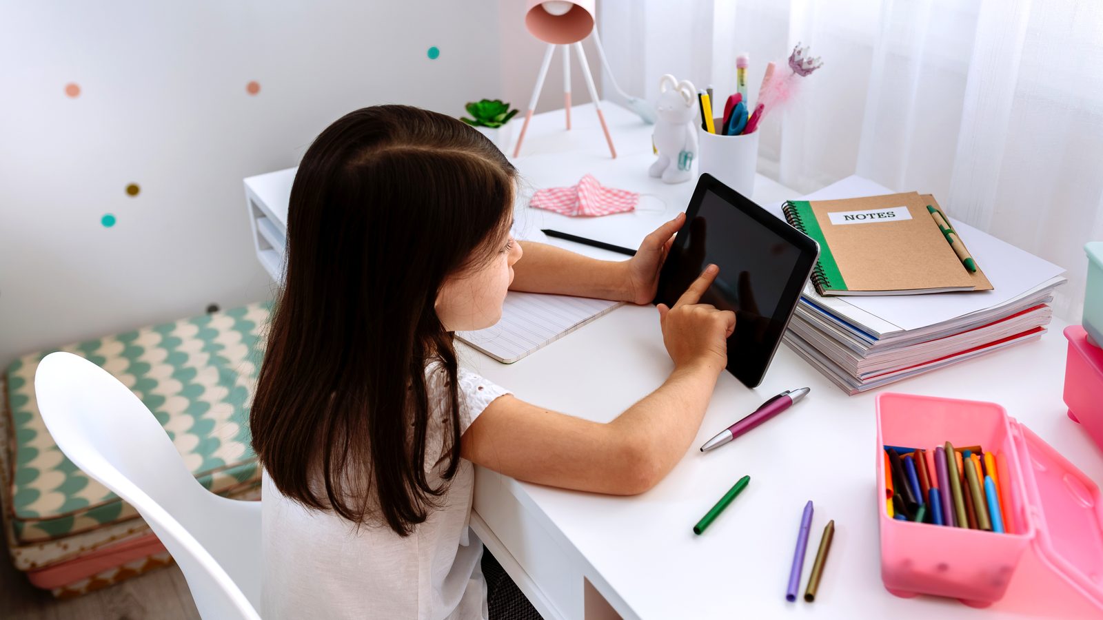 A young girl at a white desk taps a tablet, surrounded by notebooks, pens and a box of crayons.