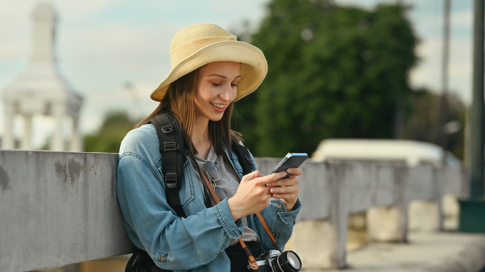A smiling woman in a sun hat and denim jacket checks her phone outdoors, with a camera hanging from her shoulder.