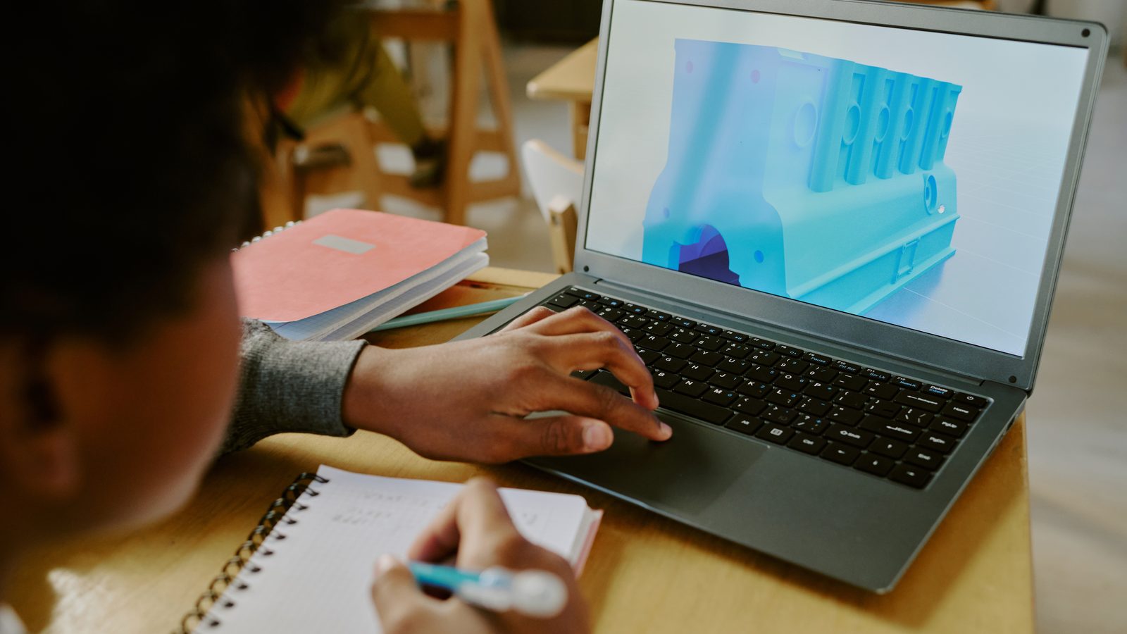 A student at a classroom desk takes notes in a spiral notebook while working on a laptop that shows a blue 3D model of a machine part.