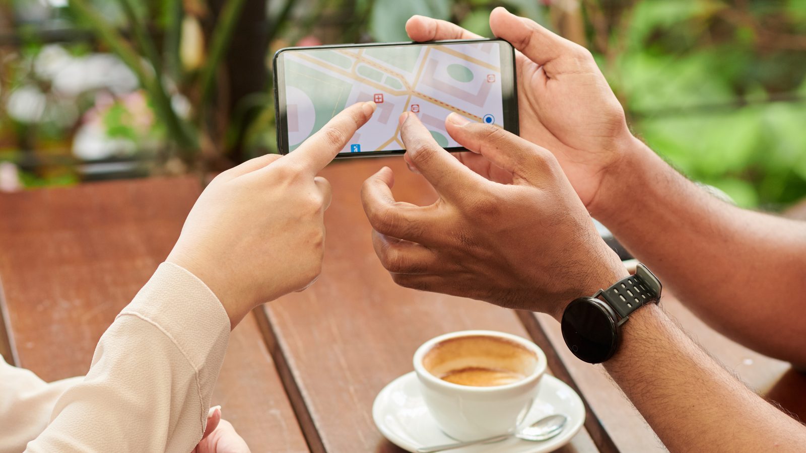 Two people at an outdoor café table point at a map on a phone next to a cup of coffee.