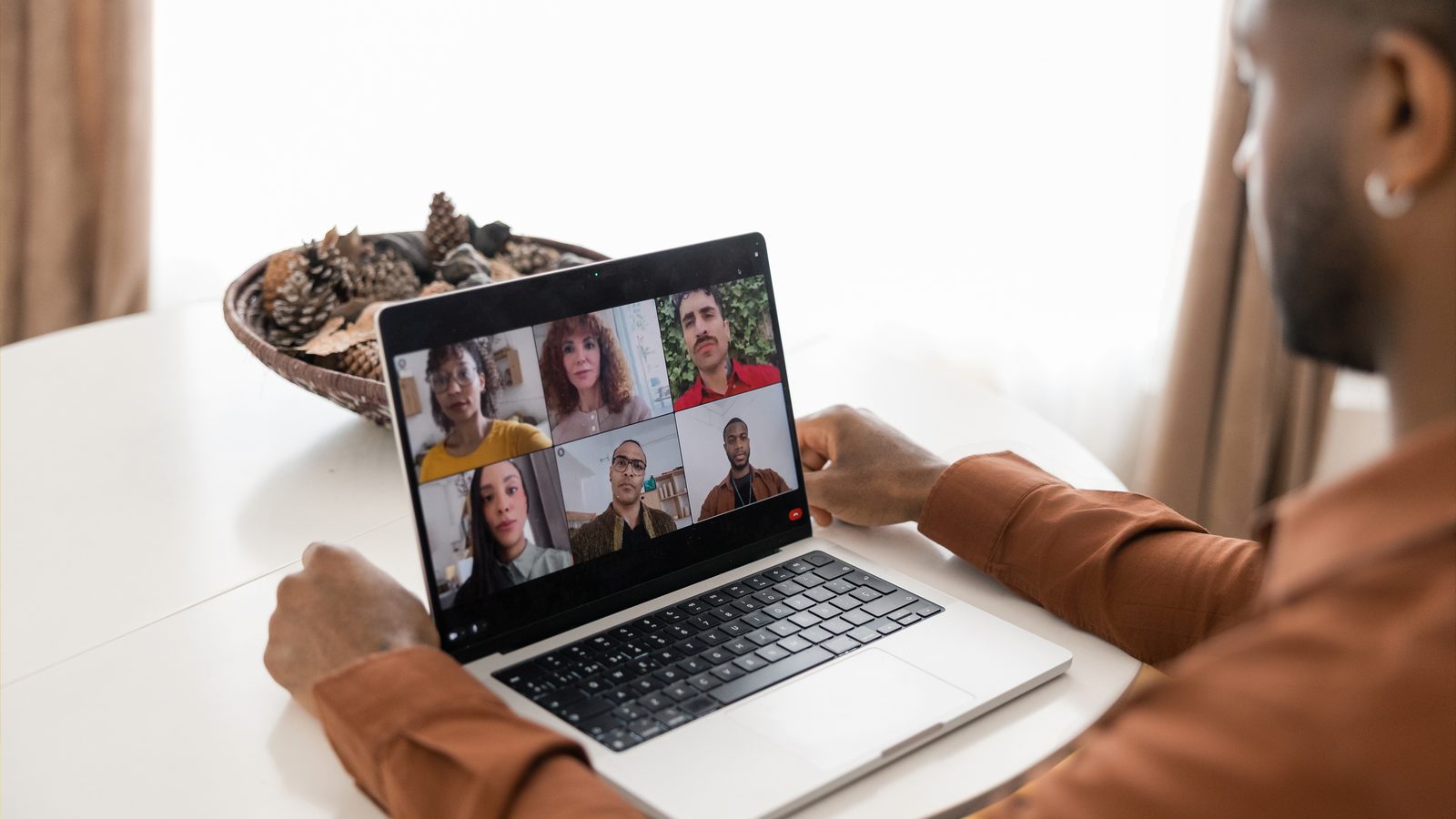 A man at home sits at a round table on a video call with six people on his laptop.
