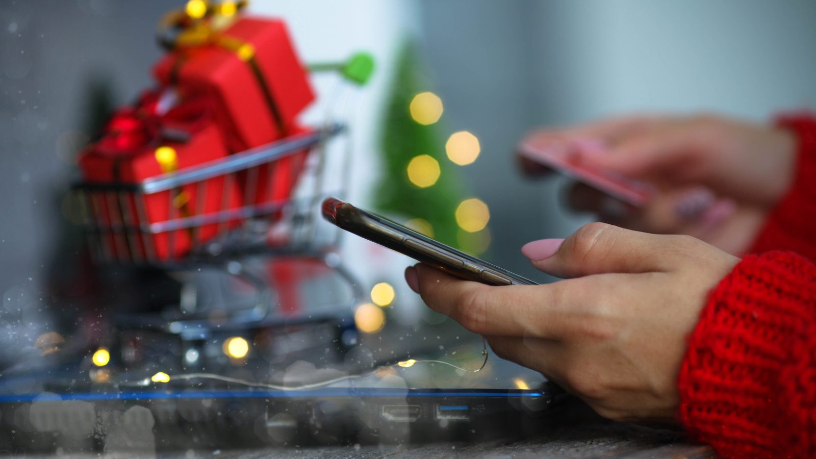 Hands in a red sweater hold a smartphone next to a tiny shopping cart full of wrapped gifts and holiday lights.