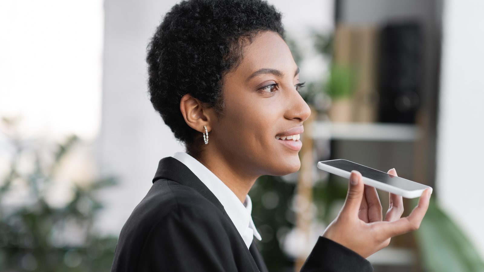 A smiling woman in a black blazer holds a smartphone flat near her mouth and talks into it in an office.