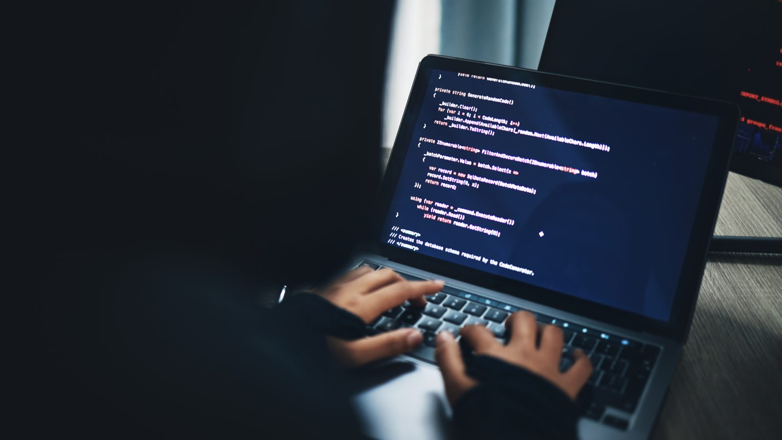 Hands type on a laptop in a dark room, its screen full of colorful lines of code, with a second monitor glowing in the background.