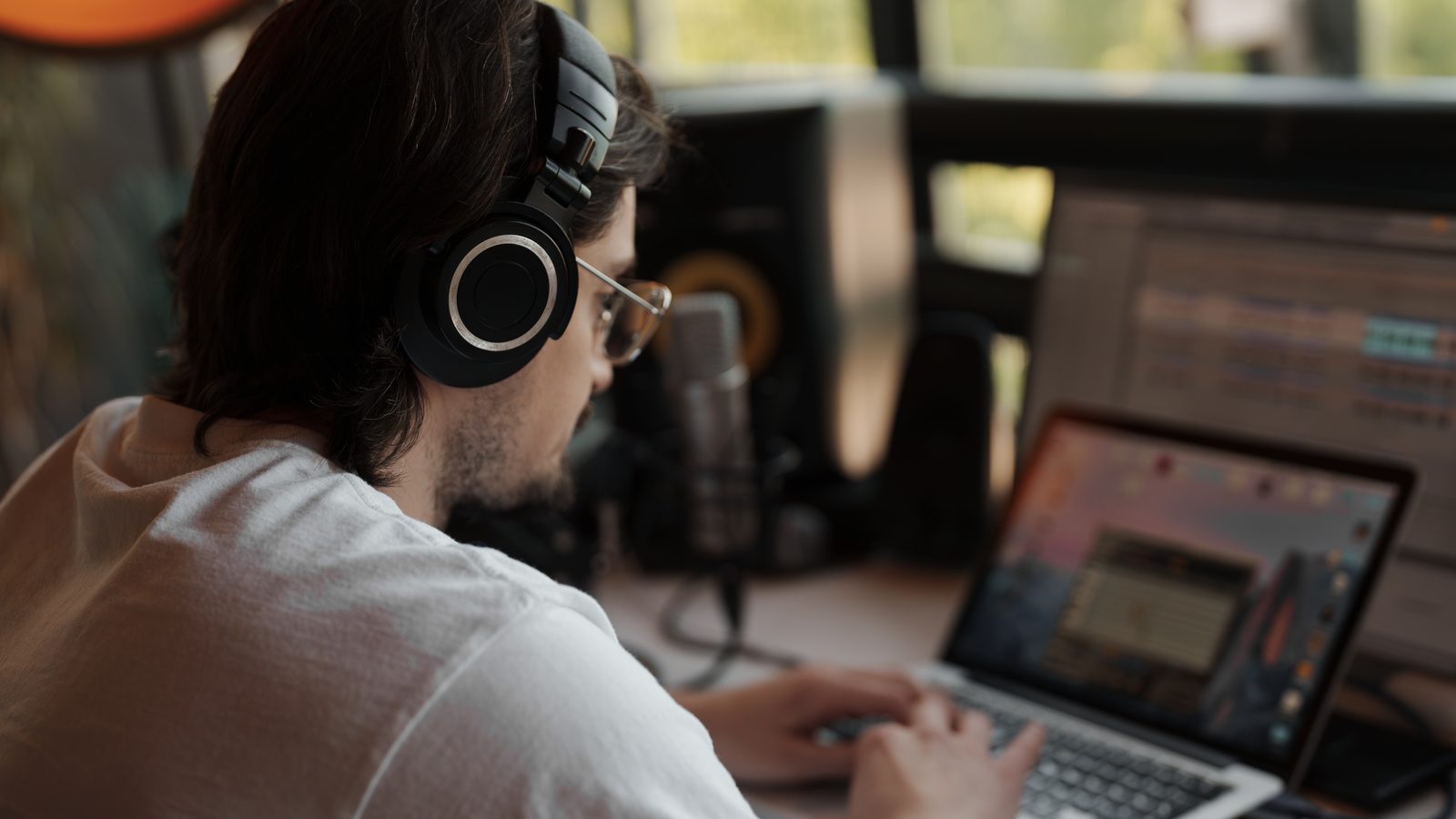 A man with glasses and headphones types on a laptop at a desk with a microphone and studio speakers.