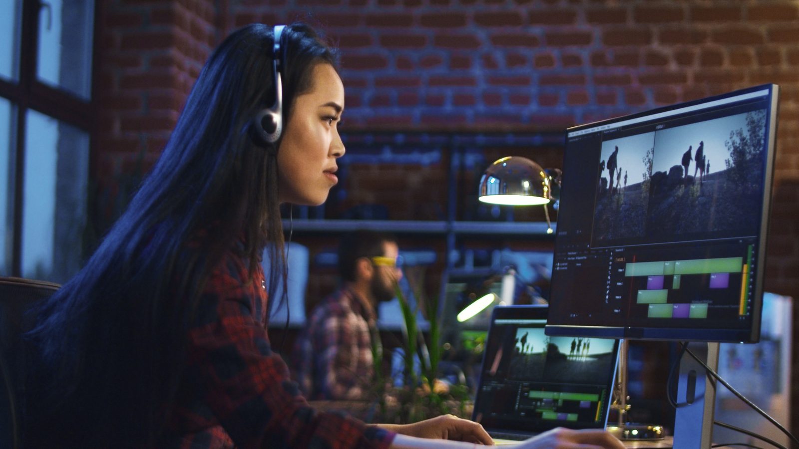 A woman in headphones edits video on a large monitor in an office at night.