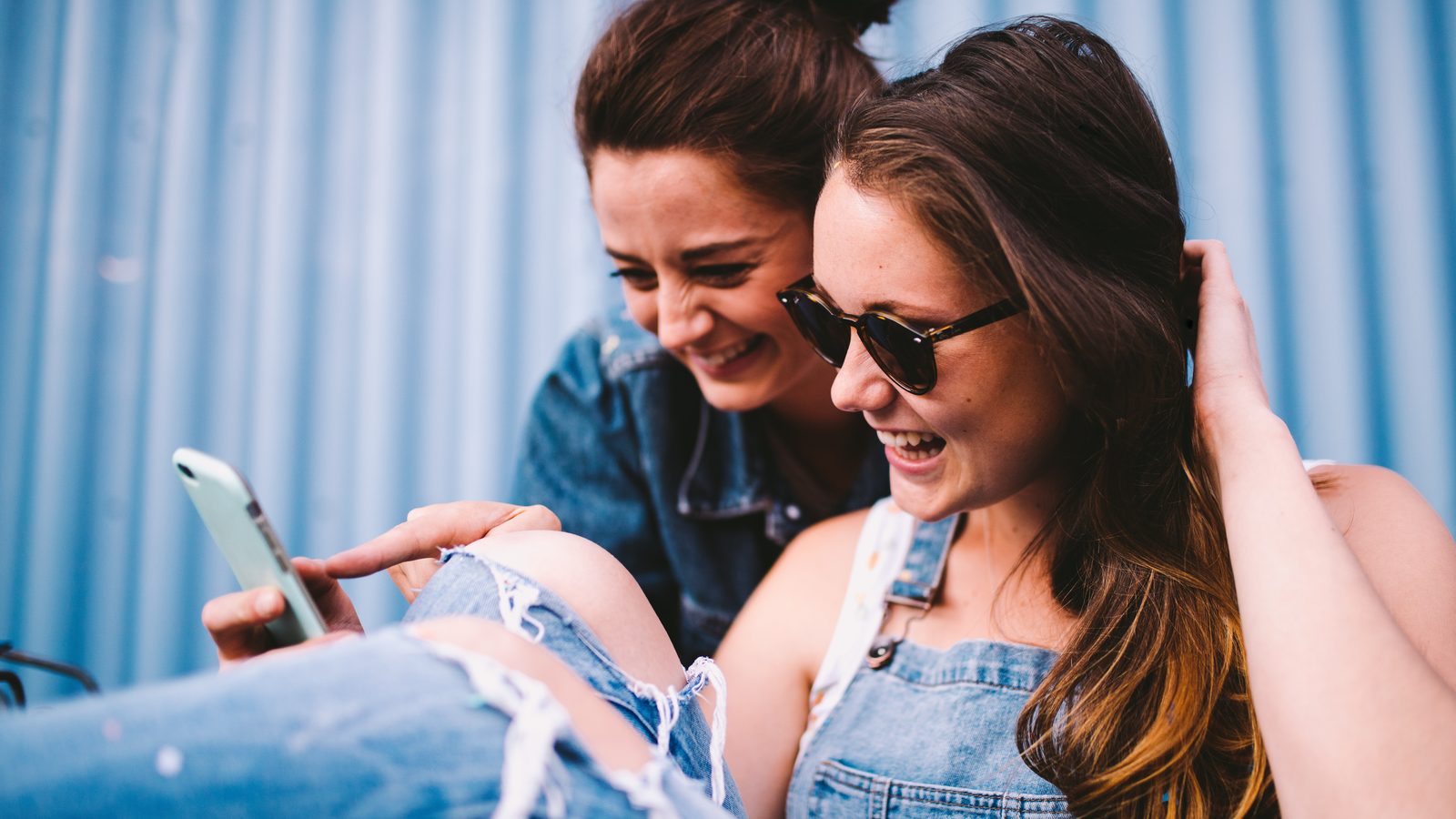 Two young women laugh while looking at a phone together.