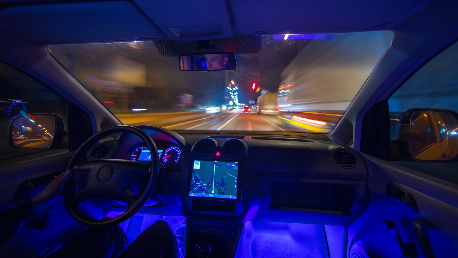 The view from inside a car driving down a city street at night, with blue cabin lighting and a map on the dashboard screen.