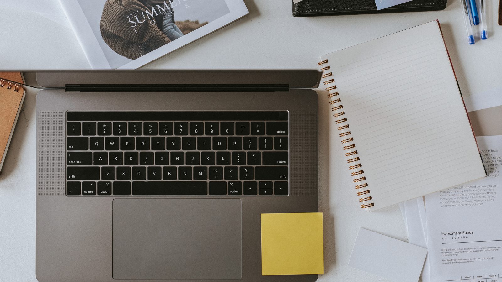 Seen from above, an open silver laptop sits on a white desk among a spiral notebook, a yellow sticky note, pens and printed papers.