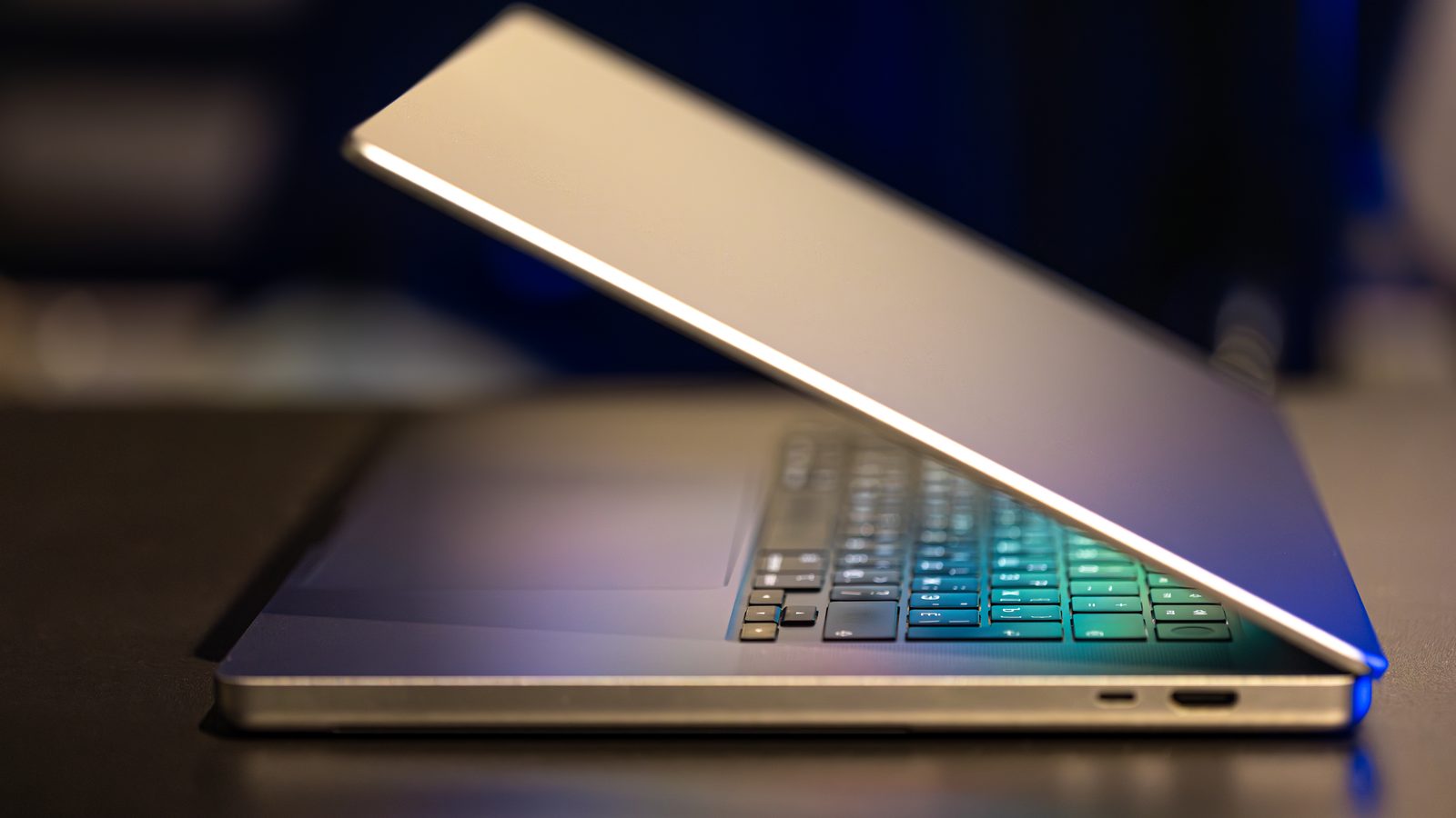 A slim silver laptop sits half open on a dark desk, its keyboard glowing in blue and green light.