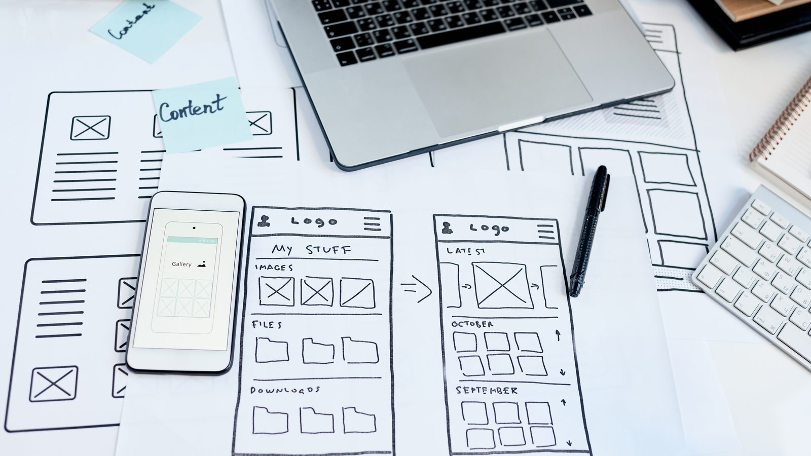 Hand-drawn wireframes of phone app screens are spread across a desk beside a smartphone, a laptop, a keyboard and a black pen.