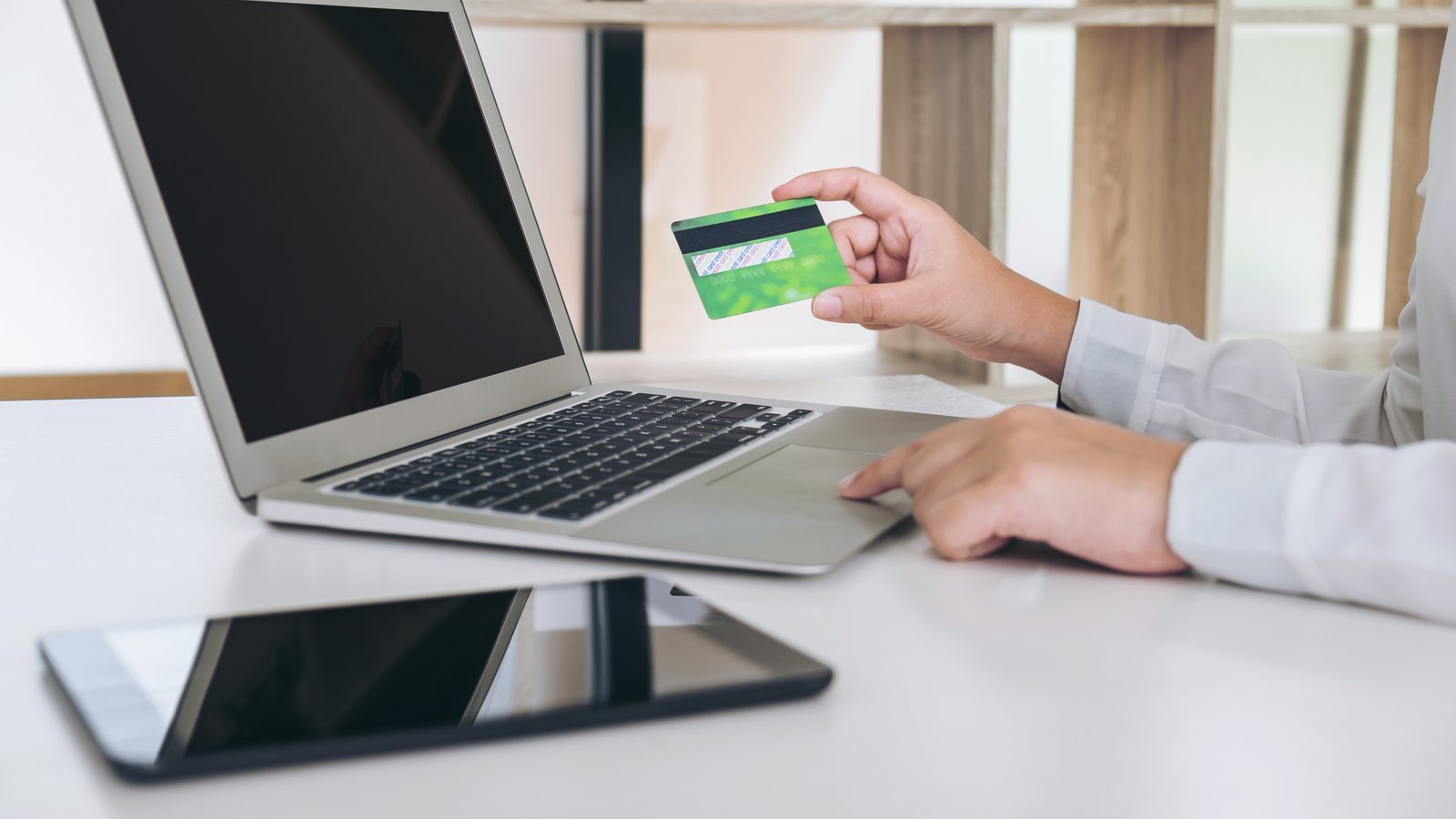 A person holds a green payment card while typing on a laptop, with a tablet lying on the white desk in front.