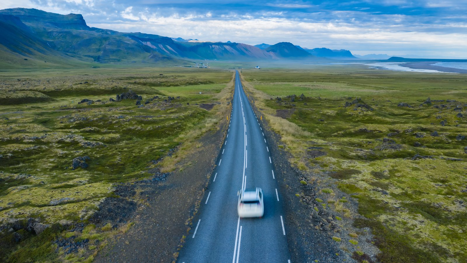 A white car drives down a long, empty two-lane road through green, rocky country toward distant mountains.