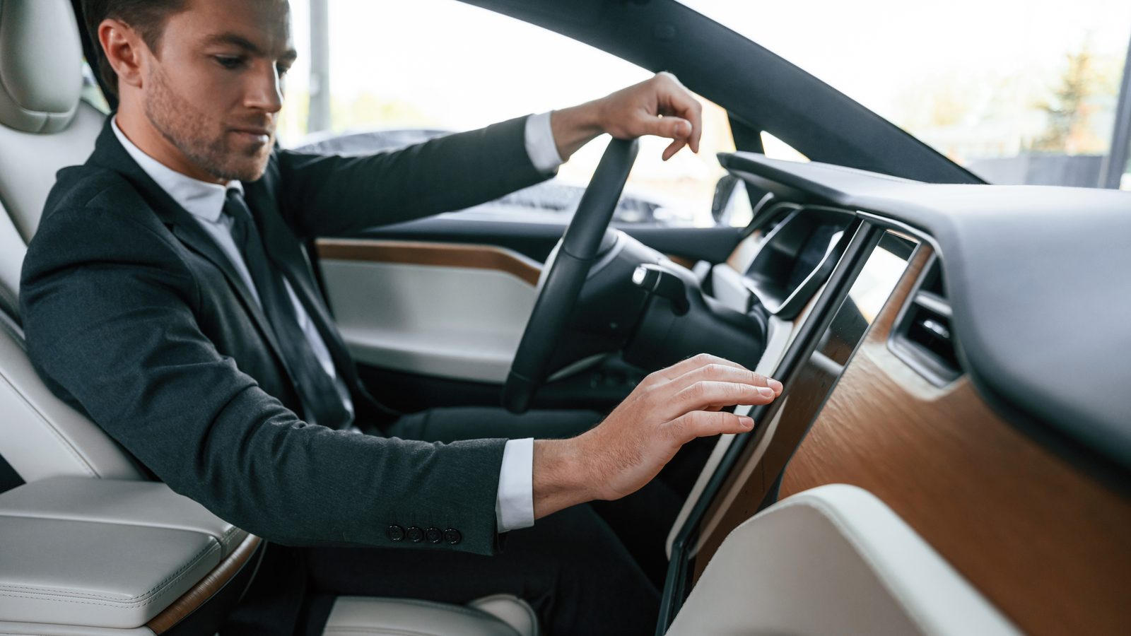 A man in a suit sits in a car's driver's seat and reaches for its large central touchscreen.