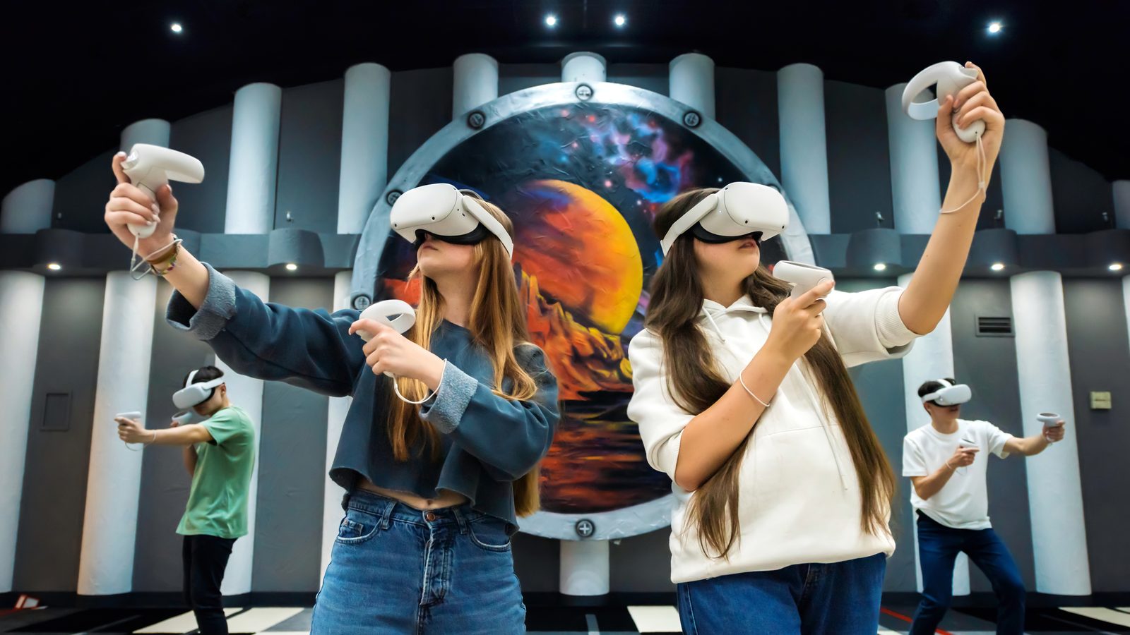Teenagers wearing white VR headsets swing hand controllers in a room with striped columns and a large painted planet on the back wall.