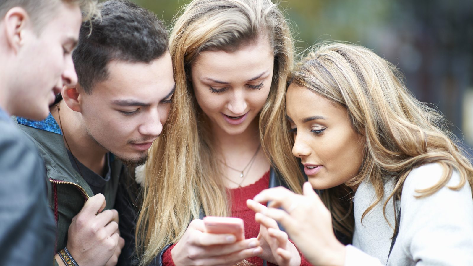 Four young friends crowd together outdoors to look at one smartphone, as one woman points at the screen.