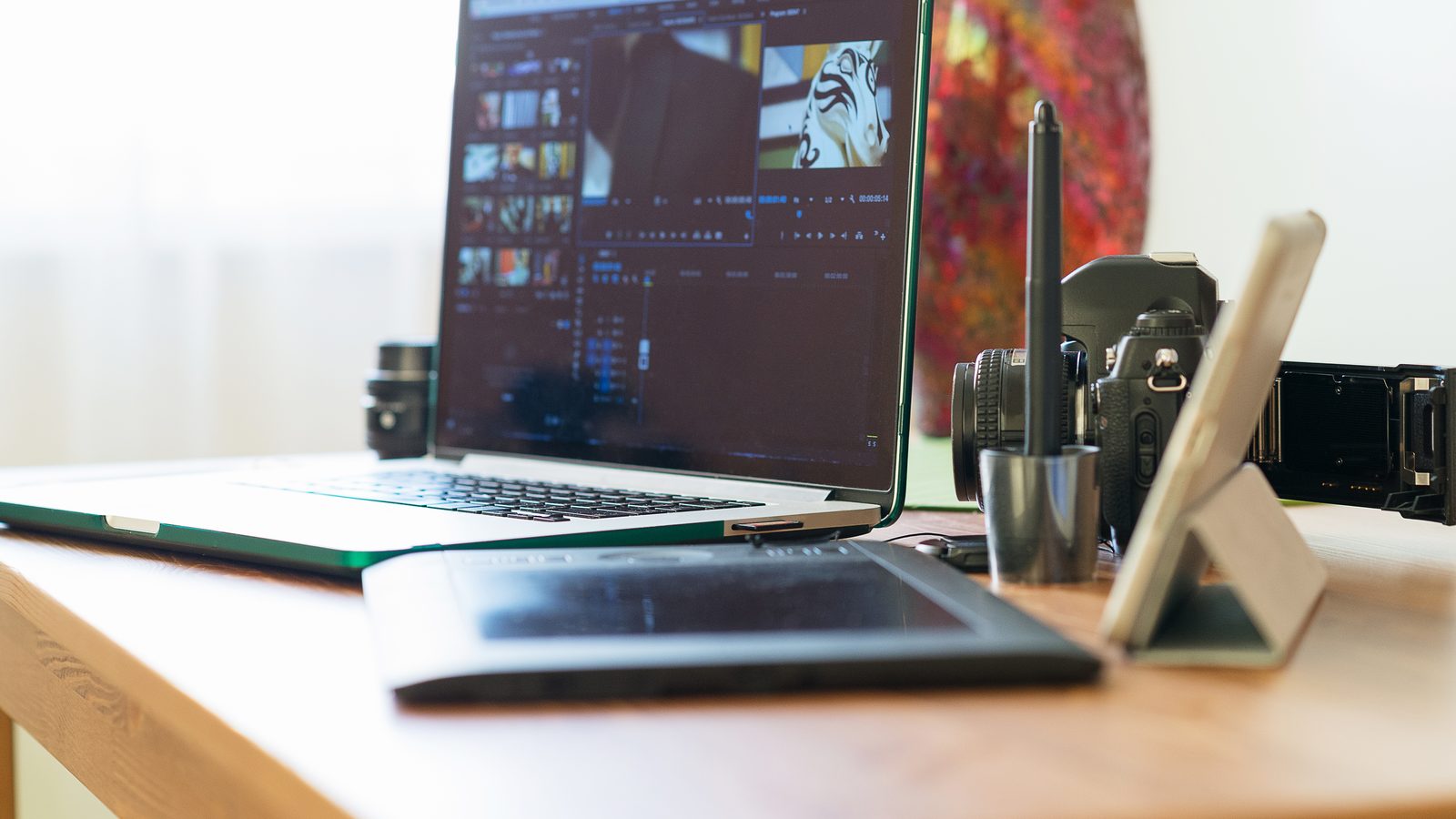 A laptop showing photo and video editing software sits on a wooden desk beside a camera, a drawing tablet and a stylus.