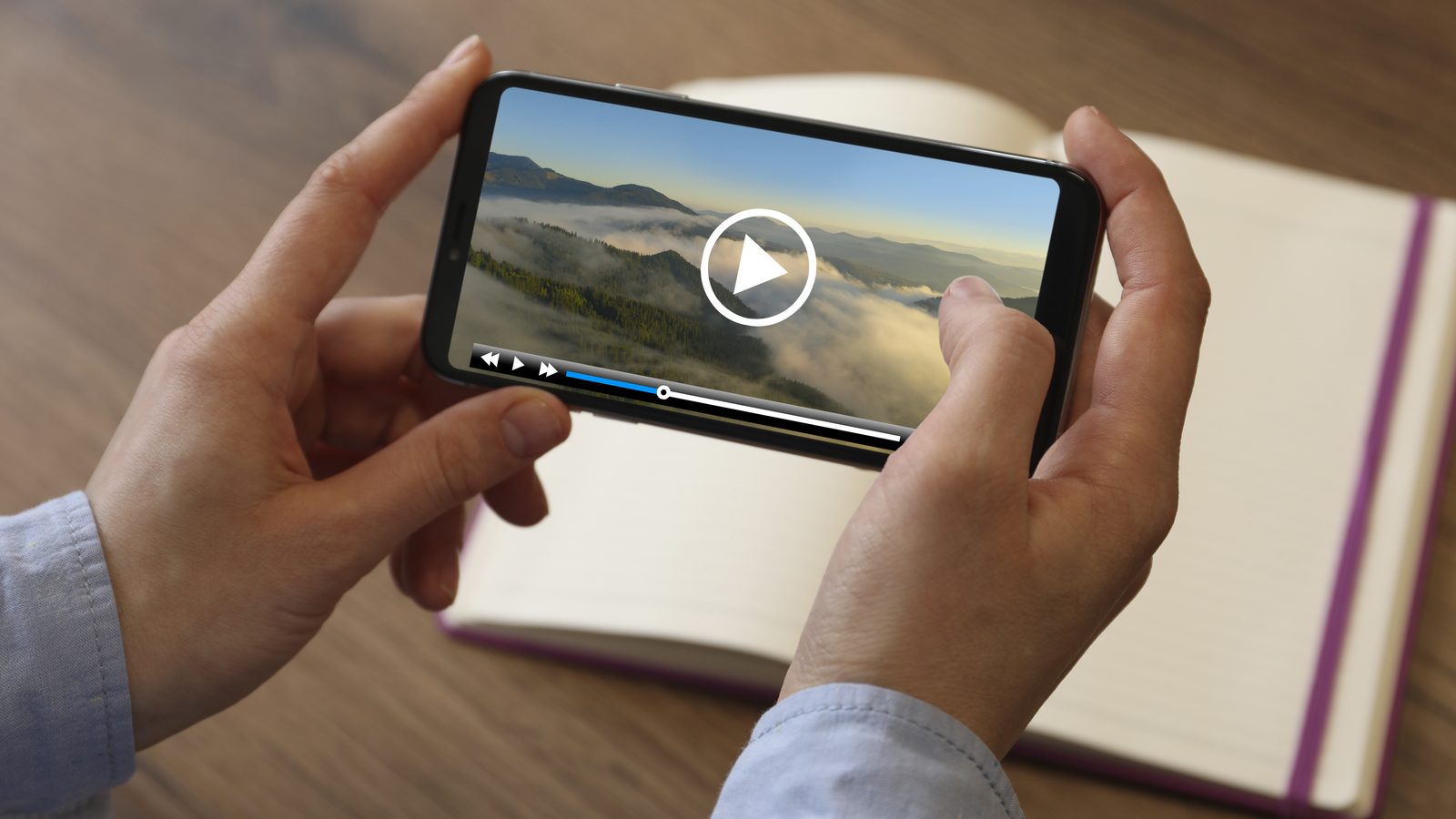 Hands holding a smartphone sideways, playing a video of mountains and clouds above an open notebook on a wooden table.